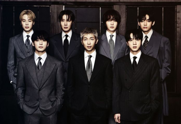 BTS dressed in stylish suits, standing together in a formal setting with a dark wooden background.