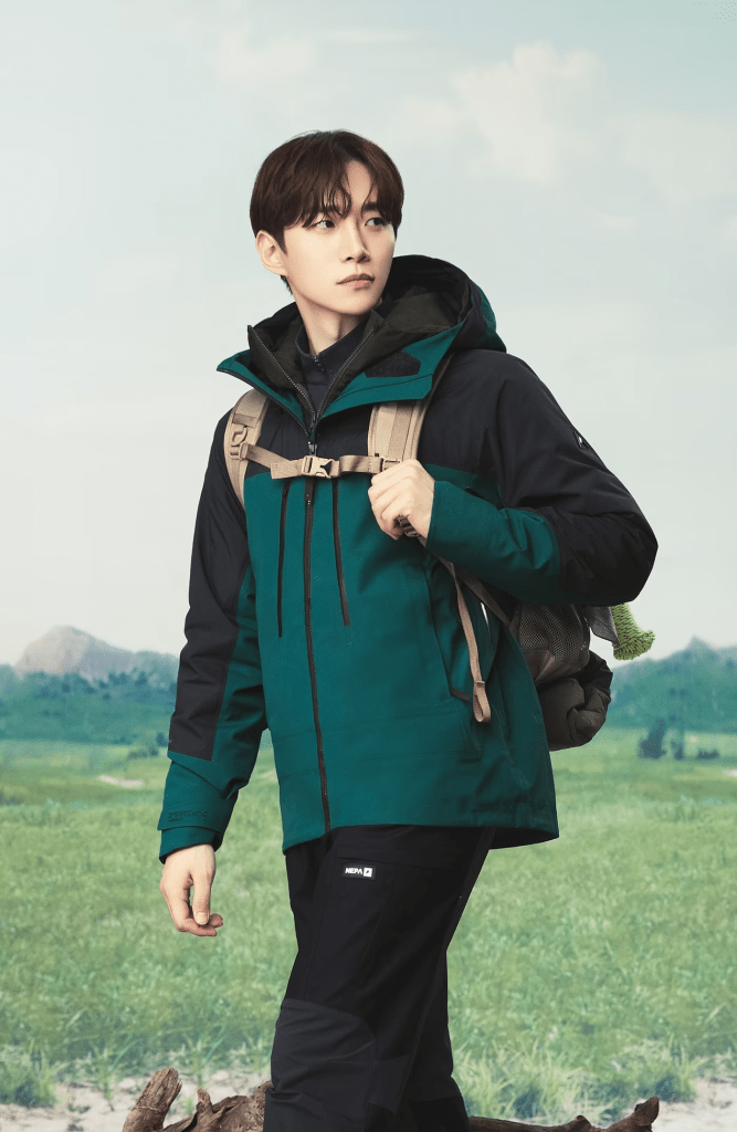 Lee Junho wearing a teal and black outdoor jacket with a backpack, standing in a grassy field with mountains in the background.