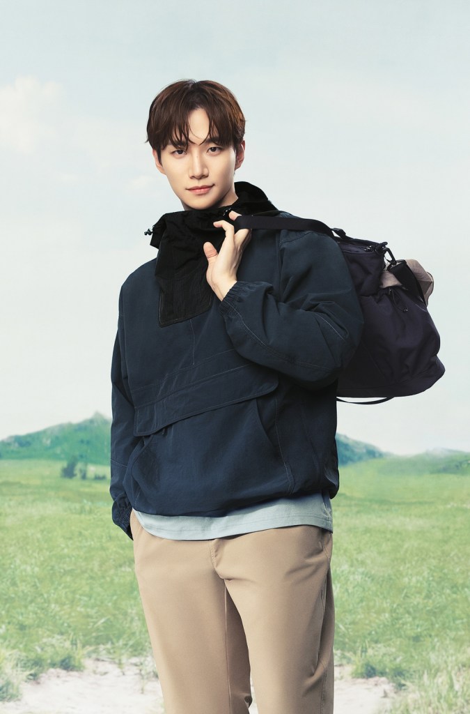Lee Junho wearing a navy blue jacket and light beige pants stands outdoors, smiling while holding a backpack over one shoulder, with a grassy landscape and blue sky in the background.