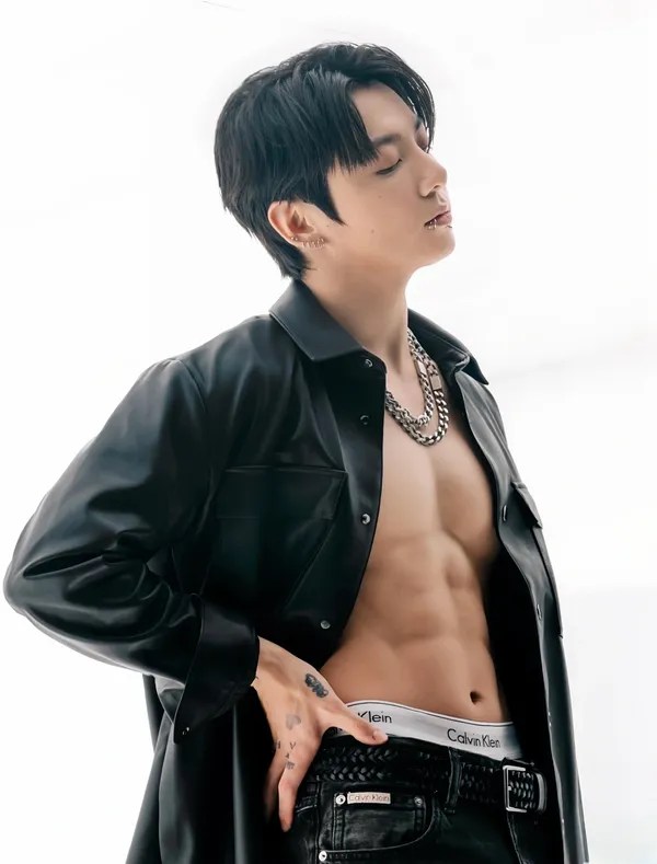 Jungkook in a black leather shirt poses confidently, showing off his toned abs and wearing a chain necklace. He stands with his eyes closed against a light background.