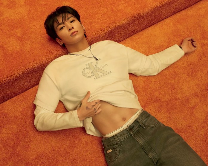 Jungkook reclines on a vibrant orange carpet, wearing a white Calvin Klein long-sleeve shirt with the brand logo visible. He has a piercing and tattoos, with a relaxed expression as he poses with one hand on his stomach and the other extended beside him.