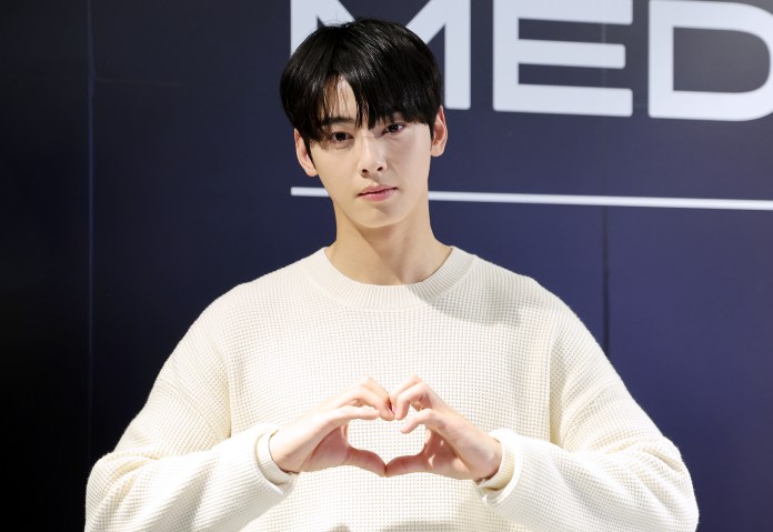 Cha Eun-woo in a cream-colored sweater making a heart shape with his hands, standing in front of a backdrop with the word 'MEDIA.'