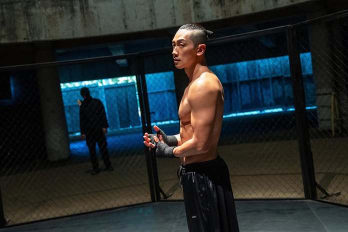 A shirtless Rain in fighter stands inside a cage in an underground arena, with a focused expression on his face. He wears hand wraps and black shorts, with a shadowy figure visible in the background.