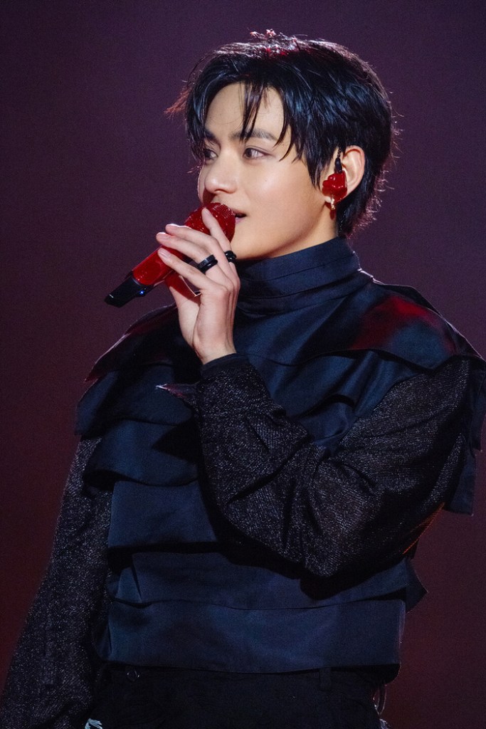 V on stage, wearing a black outfit with ruffled details, holding a red microphone and showcasing a stylish hairstyle against a blurred background.