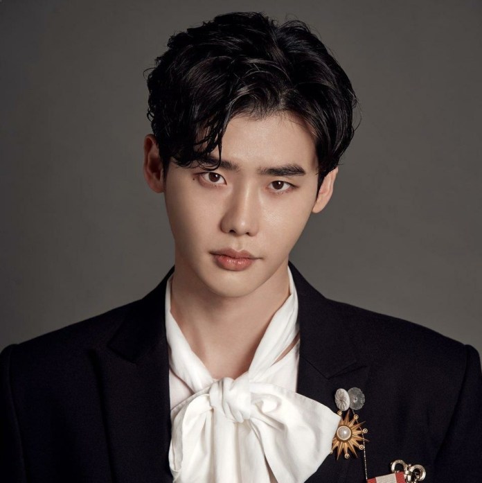 Lee Jong Suk with dark, styled hair, wearing a black suit jacket with decorative pins and a white scarf tie, looking directly at the camera.