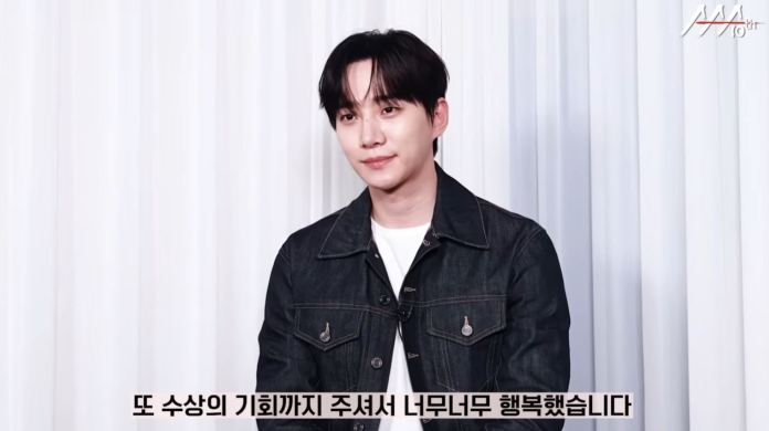 Junho wearing a denim jacket and a white t-shirt, set against a white curtain backdrop, smiling softly while seated.