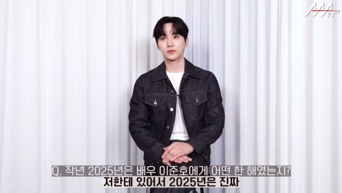 Junho seated in front of a white curtain, wearing a dark denim jacket over a white shirt, looking directly at the camera.