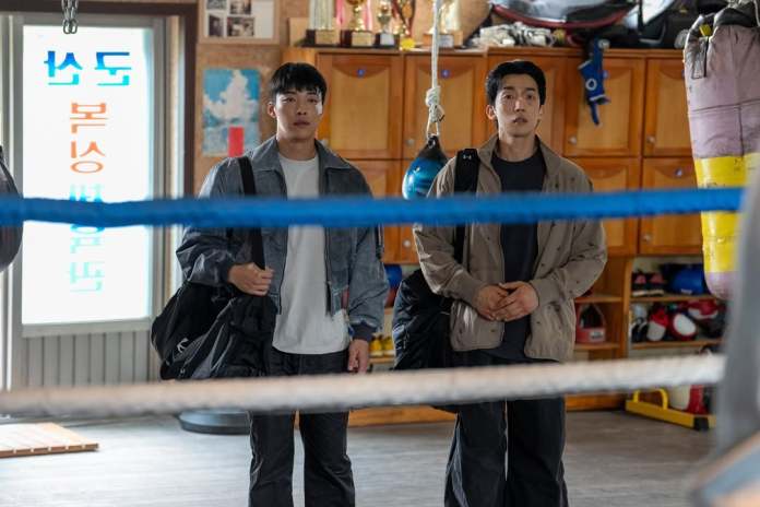 Woo Do-hwan and Lee Sang-yi standing inside a boxing gym, both carrying sports bags. The background features various trophies and boxing equipment.