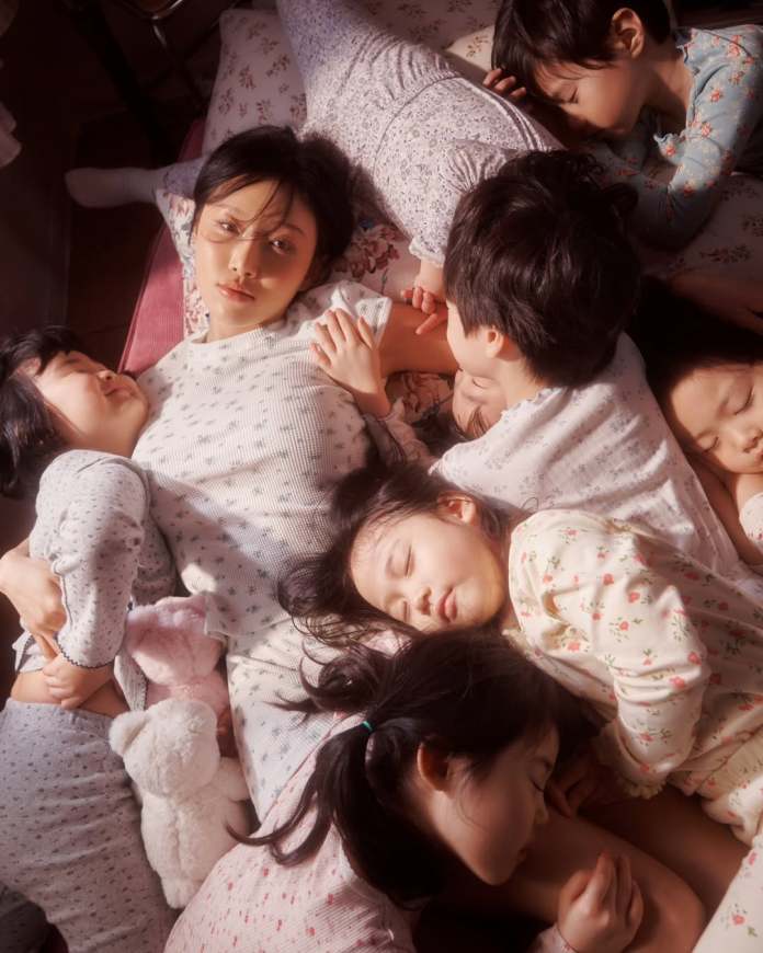 Hwasa lying on a bed surrounded by several sleeping children, all dressed in pajamas, with a soft and cozy atmosphere.