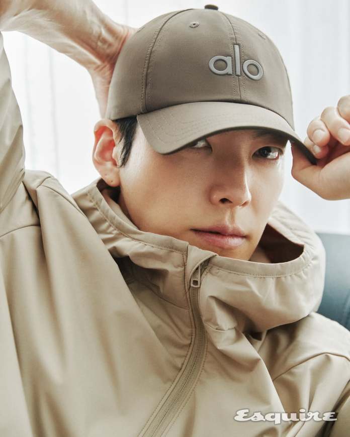 Kim Woo-bin wearing a beige jacket and a light brown cap with the logo 'alo', looking contemplatively at the camera.