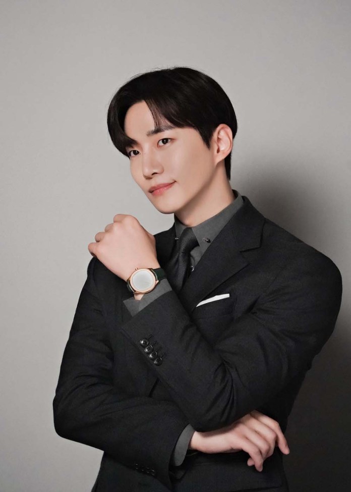 Lee Junho in a black suit posing confidently with a watch on his wrist, smiling slightly at the camera.