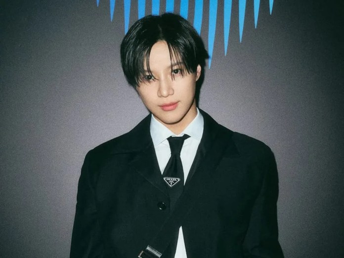 Taemin with dark hair wearing a black suit and tie, standing in front of a dark background with blue geometric patterns.
