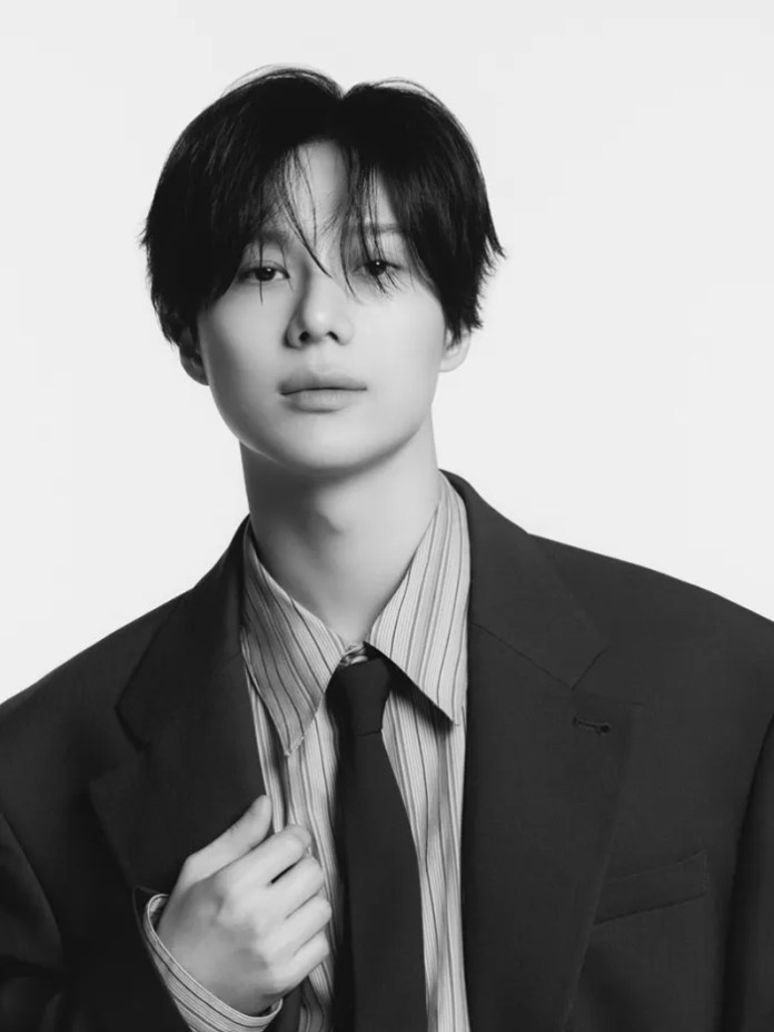 Taemin with dark hair wearing a black suit and a striped shirt, holding their tie, posed against a plain white background.