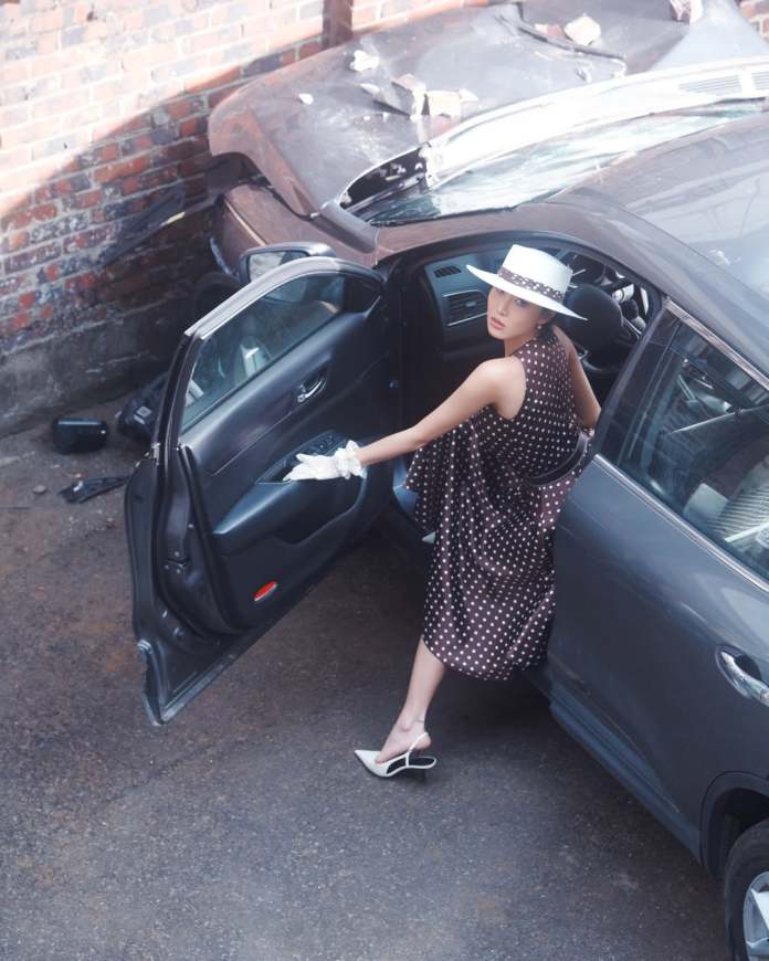 A fashionable Hwasa in a polka dot dress and white hat steps out of a car, with a damaged vehicle and brick wall in the background.