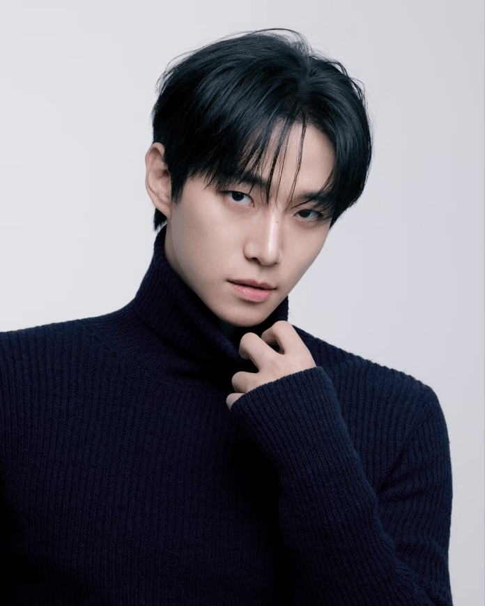 Lee Junho with dark hair poses in a close-up shot, wearing a dark turtleneck sweater. He is looking directly at the camera with a confident expression, his hand holding the collar of his sweater.