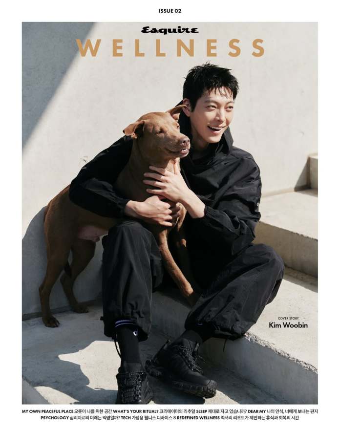 Kim Woo-bin wearing casual black clothing sits with a brown dog on his lap, smiling and enjoying the moment. The image features the words 'Esquire WELLNESS' above and has a clean, modern aesthetic.
