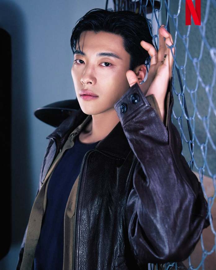 Woo Do-hwan with styled dark hair poses against a chain-link fence, wearing a black leather jacket over a casual shirt.