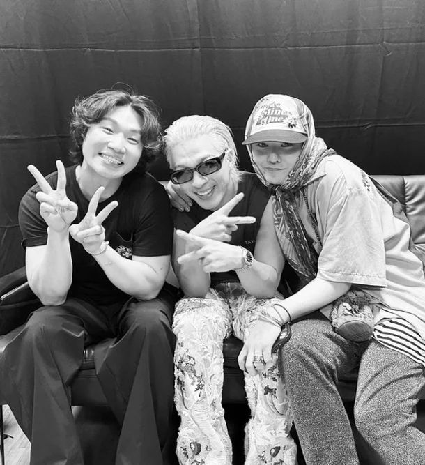 BIGBANG posing together with peace signs in a black and white photo. They are sitting on a couch with a black backdrop.