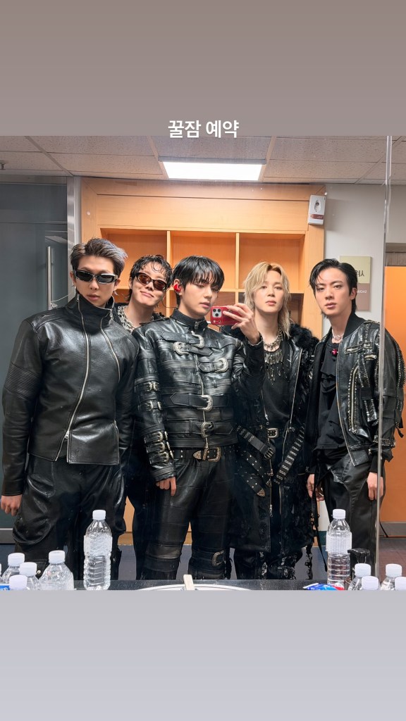 BTS posing together in stylish black leather outfits, standing in front of a mirror with water bottles on a table in the foreground.