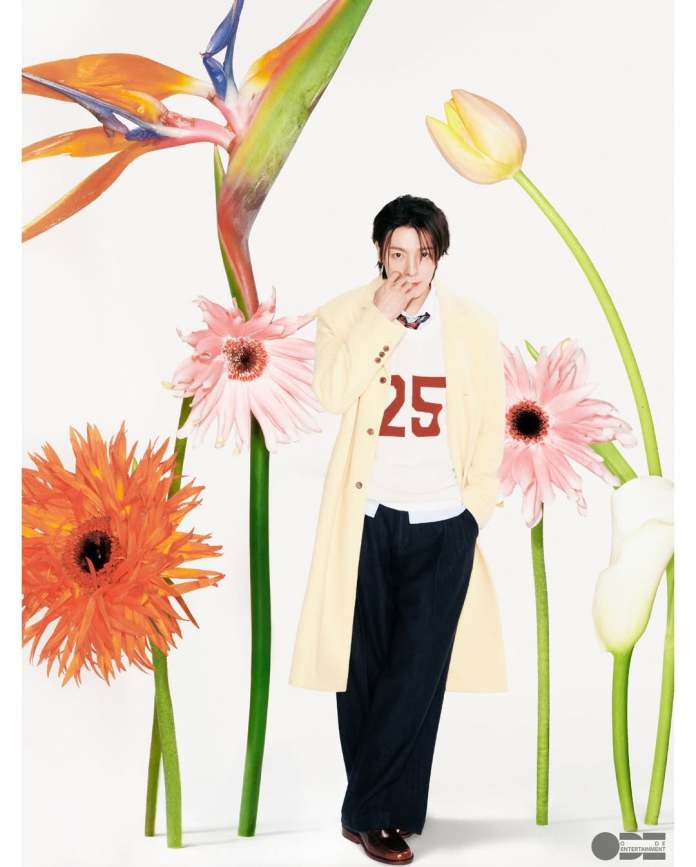 A stylish person wearing a cream-colored coat with a red number 25 on their shirt, standing among vibrant flowers including orange gerbera daisies, pink blooms, and tropical plants against a light background.