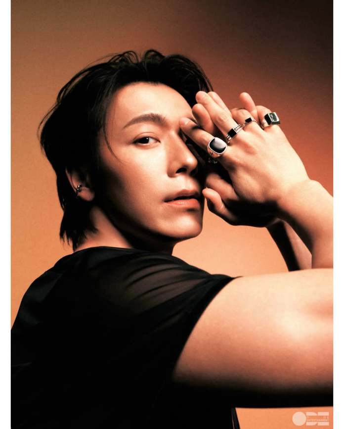 A side profile of Donghae with long dark hair, wearing a sheer black top and multiple rings, posing with his hands near his face against a warm-toned background.