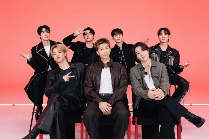 BTS posing for a photo against a vibrant pink background, dressed in stylish outfits, some sitting and others standing, showcasing a cheerful and engaging atmosphere.