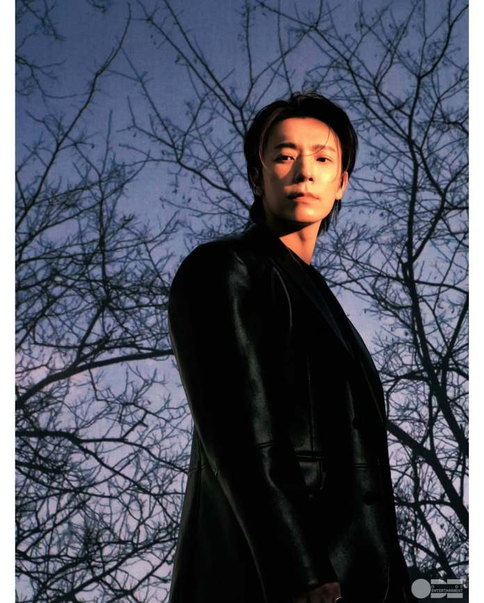 A person standing against a dark blue sky with bare tree branches, wearing a black coat and looking thoughtfully towards the camera.
