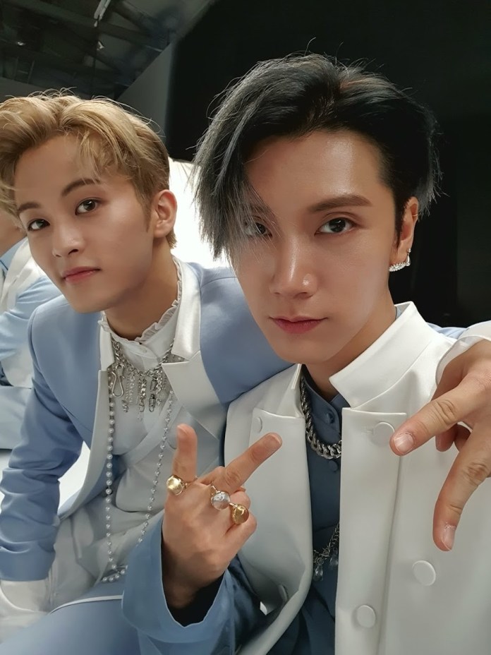 Mark and Ten posing together for a selfie, both wearing stylish outfits in light blue and white, with accessories and jewelry. The person on the left has light blonde hair, while the one on the right has dark hair and gives a peace sign.