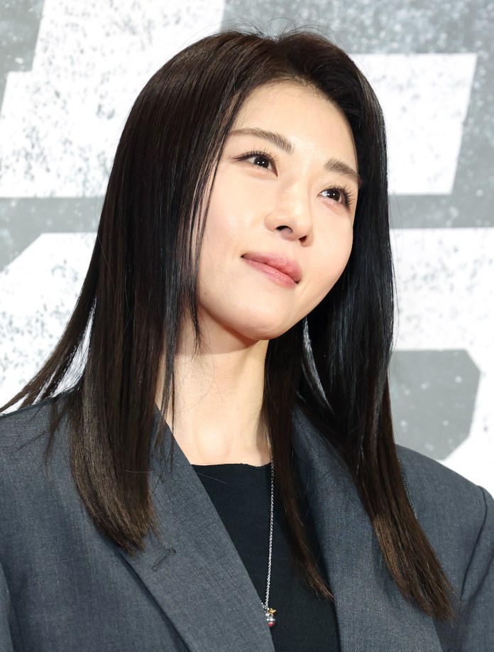 Ha Ji Won with long, straight black hair wearing a dark blazer, gazing upward with a serene expression against a textured background.