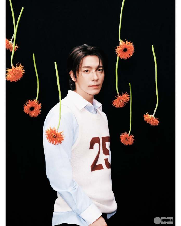 Donghae wearing a white sleeveless sweater with the number 25, posing against a black background adorned with hanging orange flowers.