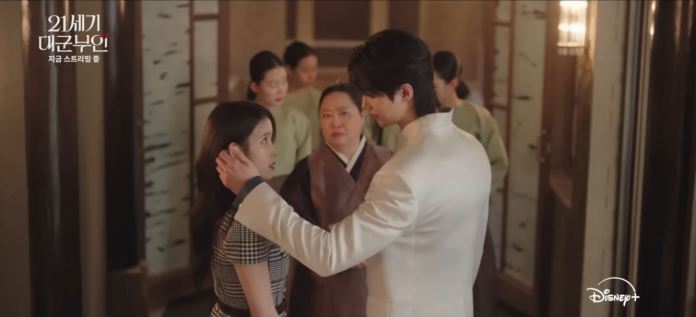 A dramatic scene featuring Byeon Woo-seok in a white suit holding the face of IU in a checkered dress, with a serious expression. In the background, two women in traditional attire observe the interaction.