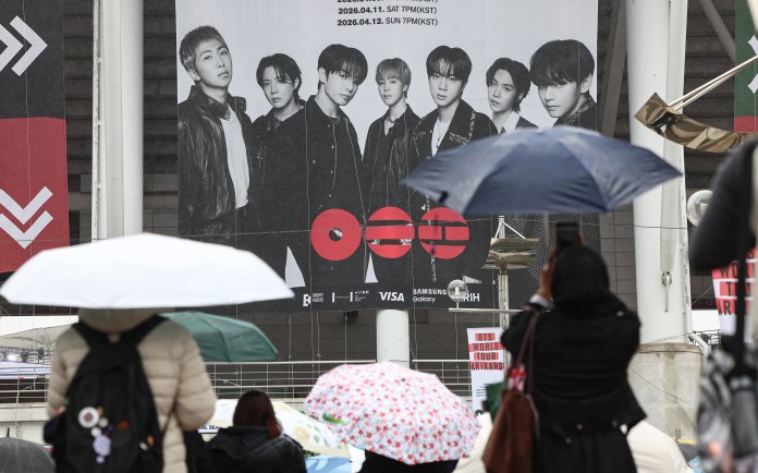 A large promotional poster featuring a group of seven male artists stands prominently above a crowd holding umbrellas. The background shows a rainy day atmosphere with people in winter clothing and an event banner for a concert.
