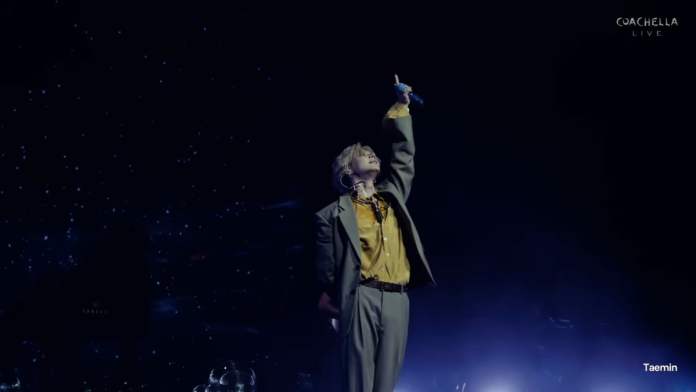 Taemin dressed in a suit raises one arm towards the sky, illuminated by stage lights, against a backdrop of stars.