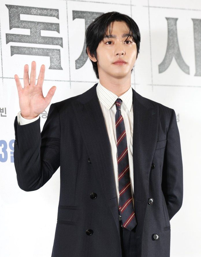 Ahn Hyo-seop in a dark suit and striped tie is standing with one hand raised in a greeting gesture.