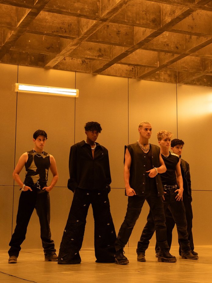 SANTOS BRAVOS members standing in a modern, illuminated room with concrete walls, showcasing diverse fashion choices.