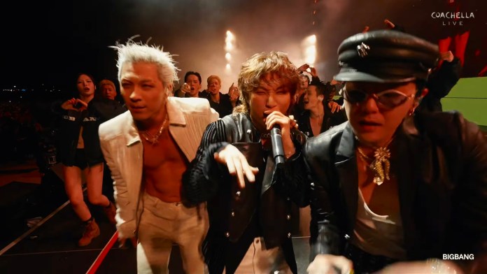 BIGBANG singing and dancing on stage at a lively outdoor concert with a large crowd in the background.
