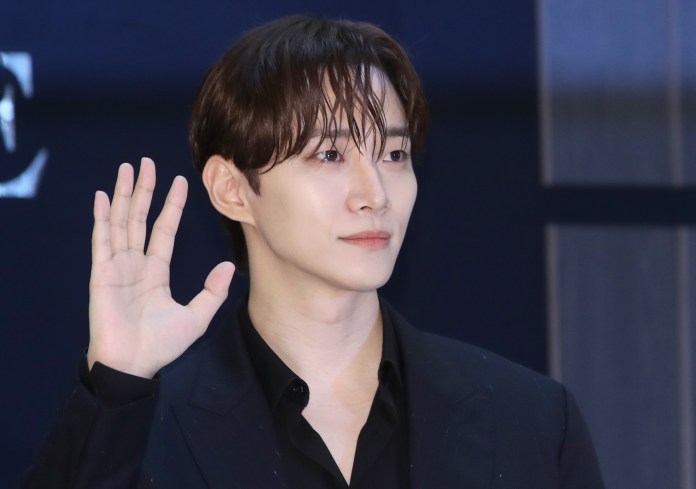 Lee Junho in a black suit waves, with slicked-back hair and a neutral expression, against a dark background.