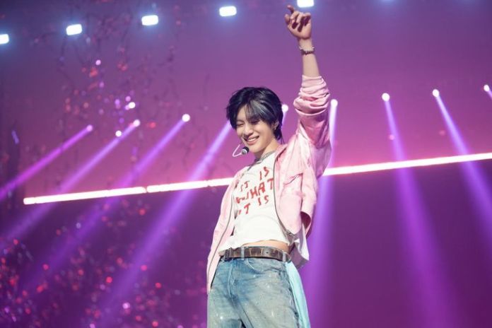 Taemin, raising one arm in excitement, wearing a pink jacket and light blue jeans, with colorful stage lights in the background.