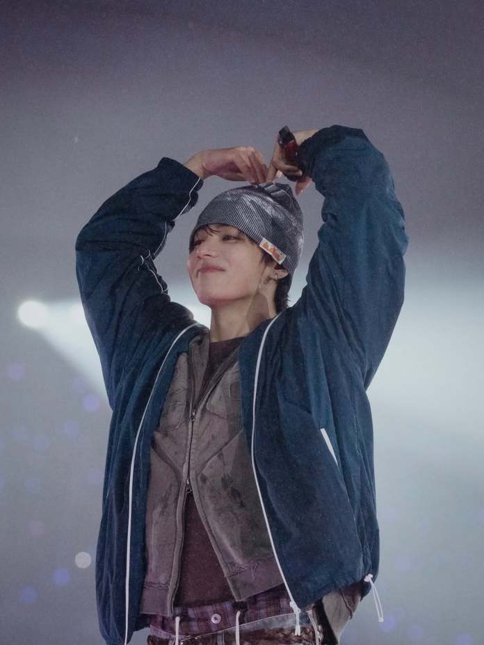 V in a stylish outfit performing on stage, wearing a beanie and a jacket, with arms raised in a joyful pose.