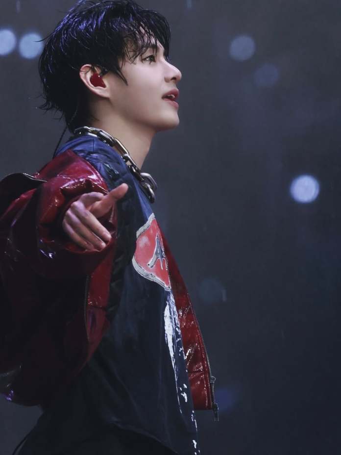 V on stage with wet hair, wearing a glossy red jacket and black shirt, reaching out with one hand and gazing upwards.