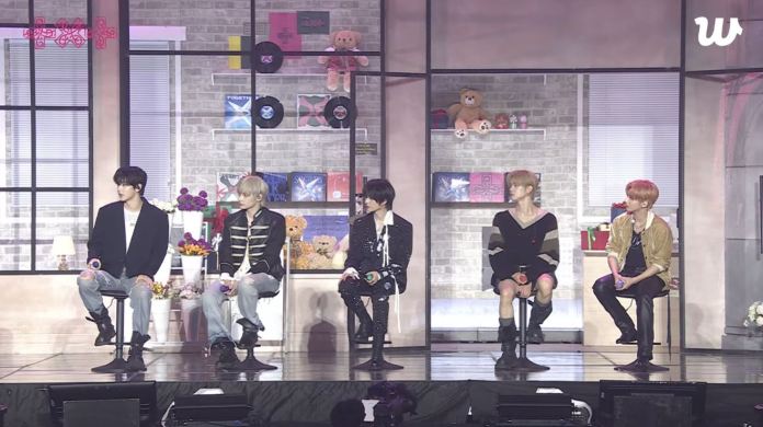 TXT members seated on stage during an event, with a colorful background featuring plush toys and decorative shelves.