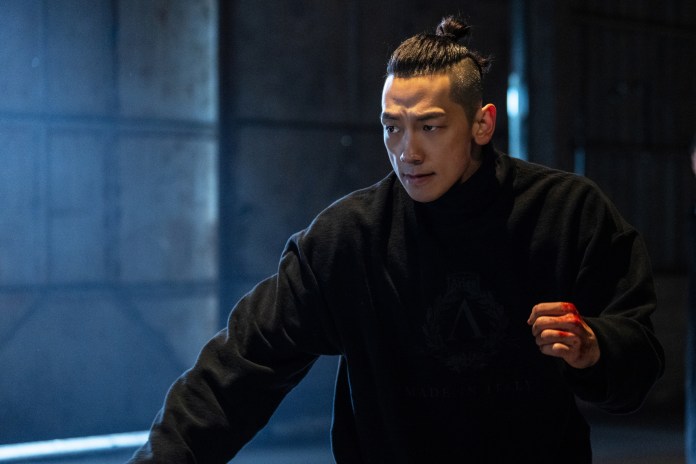 Rain with a top knot hairstyle, wearing a black oversized sweatshirt, has a focused expression while holding his right hand, which shows signs of injury, in a dimly lit industrial setting.