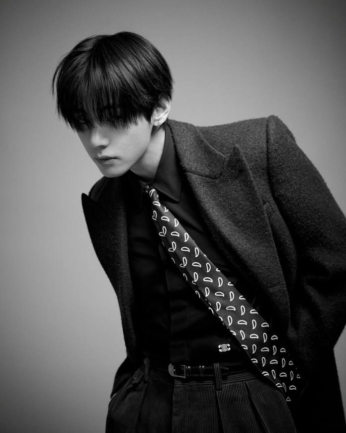 V with styled black hair, wearing a tailored coat over a black shirt and a patterned tie, gazing downwards.