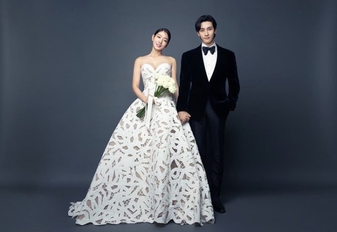 Park Shin-hye and Choi Tae-joon standing together, the woman in a detailed white wedding dress holding a bouquet of flowers, and the man in a formal black tuxedo with a bow tie.