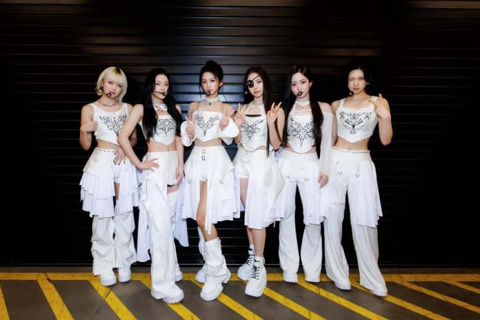 NMIXX members dressed in coordinated white outfits with intricate designs, standing in a line against a dark backdrop, posing with cheerful expressions.