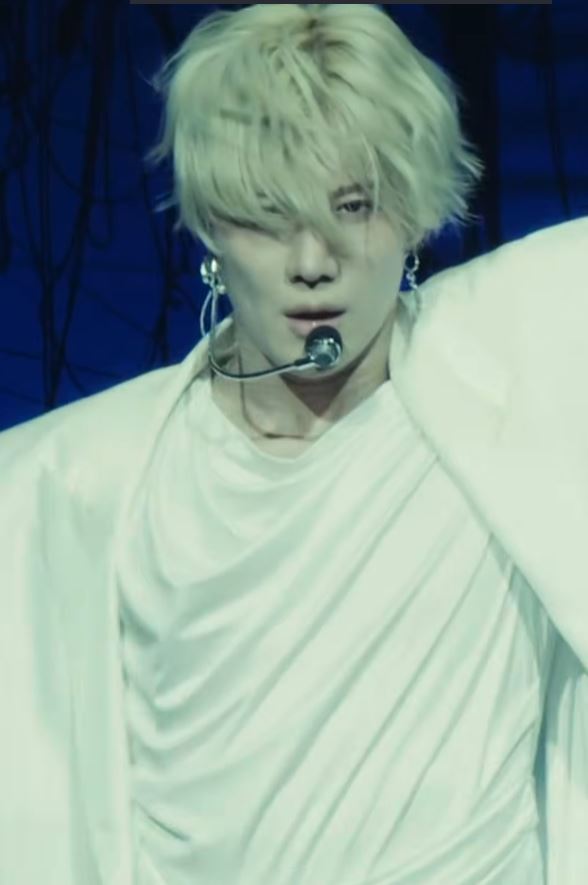 A performer with blonde hair and a microphone stands in a dramatic pose, wearing a white outfit against a dark background.