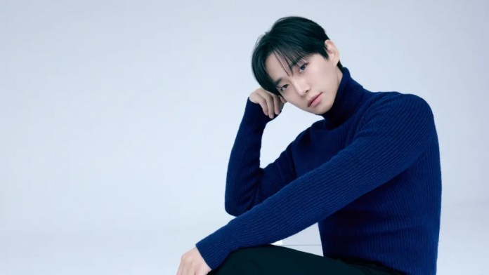 Lee Junho in a navy blue turtleneck sweater sitting thoughtfully against a pale background.