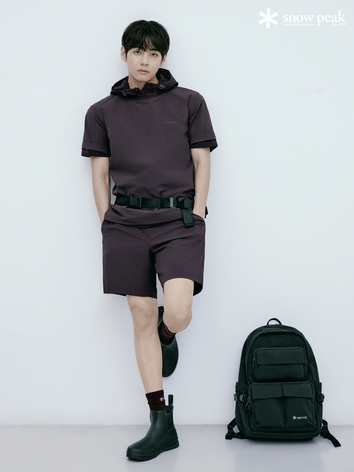 BTS's V dressed in a short-sleeved purple outfit and black rubber boots, leaning against a white wall next to a black backpack.