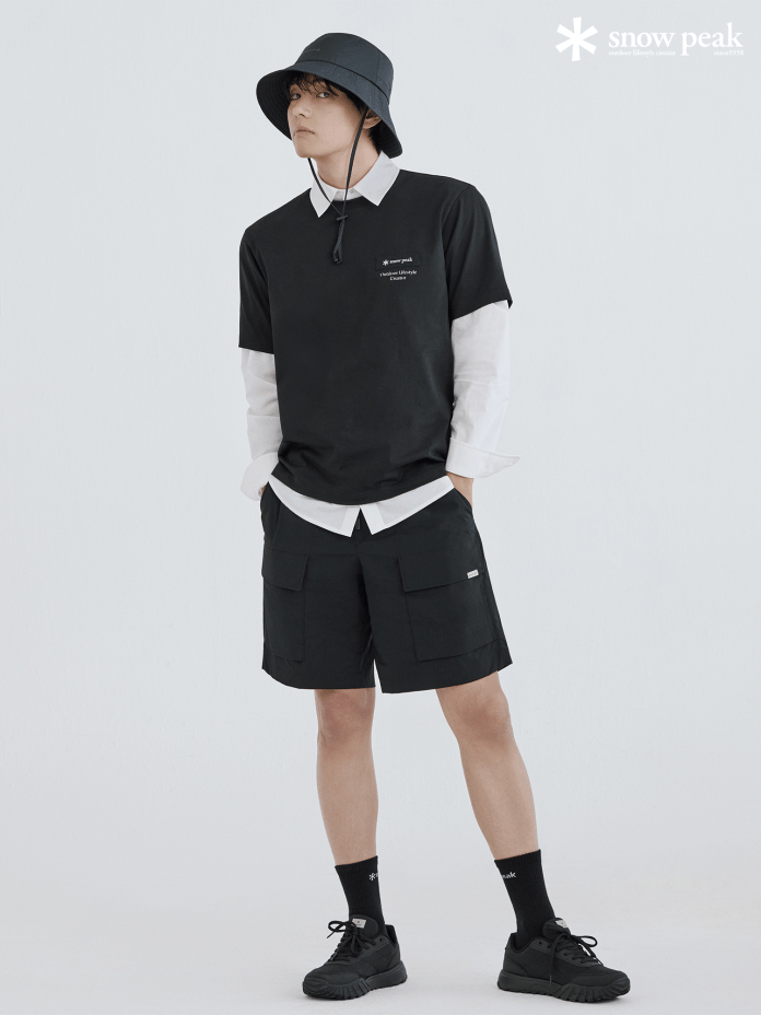 BTS's V dressed in a black t-shirt layered over a white long-sleeve shirt, wearing black shorts and a black bucket hat, standing in a neutral background.