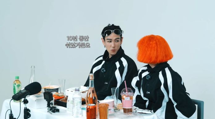 T.O.P and Nana sitting at a table with food and drinks, wearing black and white outfits. NANA has short orange hair and T.O.P has a stylish hairstyle with sunglasses.